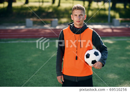Confident professional soccer coach in an orange vest and a whistle on neck at artificial football field at morning. Confident professional soccer coach in an orange vest and a whistle on neck at artificial football field at morning. 69510256