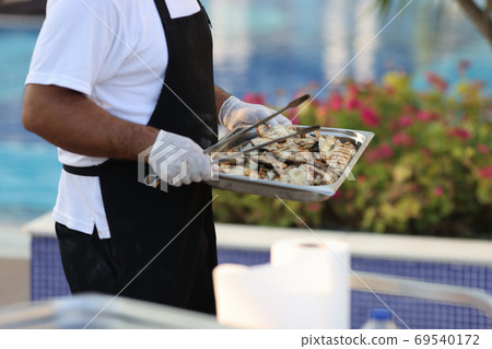 Man in black apron carries metal tray and spatula. Man in black apron carries metal tray and spatula. 69540172
