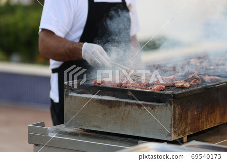 Man in black apron turn grilled meat with spatula om street. Man in black apron turn grilled meat with spatula om street. 69540173