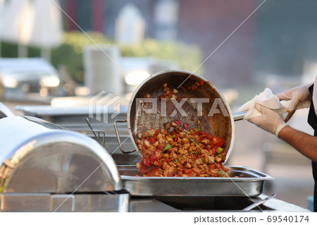 Man put cooked food from frying pan into metal container. Man put cooked food from frying pan into metal container. 69540174