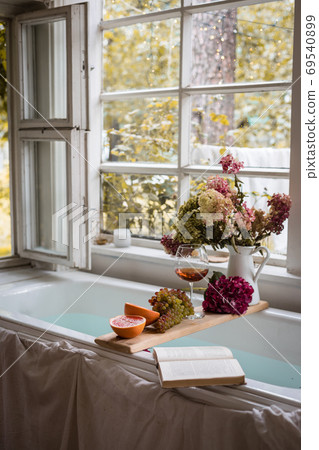 Bath tub with a tray with grapefruit slices, bunch of grapes, a glass of wine and a book 69540899