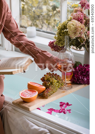 Bath tub with flower petals, grapefruit slices, bunch of grapes and woman's hand holding a glass of wine and a book 69540900