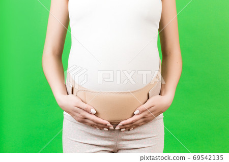 Close up of pregnant woman wearing pregnancy corset against backpain at green background with copy space. Orthopedic abdominal support belt concept Close up of pregnant woman wearing pregnancy corset against backpain at green background with copy space. Orthopedic abdominal support belt concept 69542135