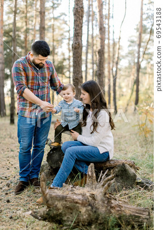 Family walk in the autumn forest. Pretty young mother and her little son sitting on an old log of a fallen tree and having fun with their handsome bearded father. 69543188