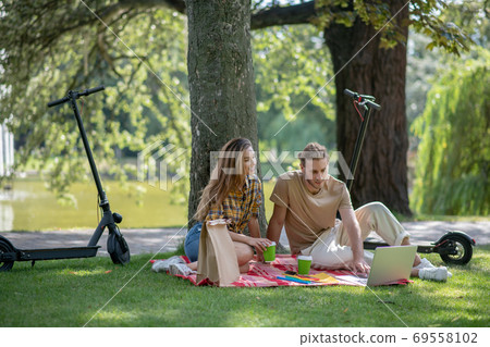 Two young people sitting under the tree and watching something on laptop 69558102