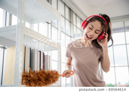 Young asian woman with headphones using a feather duster to clean wood book shelves in home . 69564614