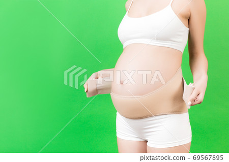 Cropped image of pregnant woman in underwear dressing orthopedic bandage to reduce pain in the back at green background with copy space. Orthopedic abdominal support belt concept Cropped image of pregnant woman in underwear dressing orthopedic bandage to reduce pain in the back at green background with copy space. Orthopedic abdominal support belt concept 69567895