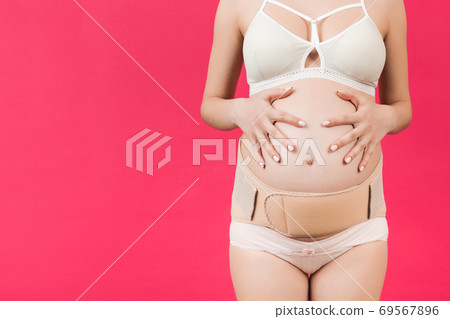 Cropped image of pregnant woman in underwear wearing elastic bandage against pain in the back at pink background with copy space. Orthopedic abdominal support belt concept Cropped image of pregnant woman in underwear wearing elastic bandage against pain in the back at pink background with copy space. Orthopedic abdominal support belt concept 69567896
