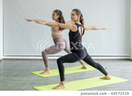Two caucasian women exercise doing yoga warrior pose stretching at home Two caucasian women exercise doing yoga warrior pose stretching at home 69572539