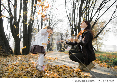 Happy young mother with her little 2 year old baby girl walking in autumn park. Pretty mom throwing up yellow autumn leaves, while her cute daughter having fun , jumping and dancing 69573206