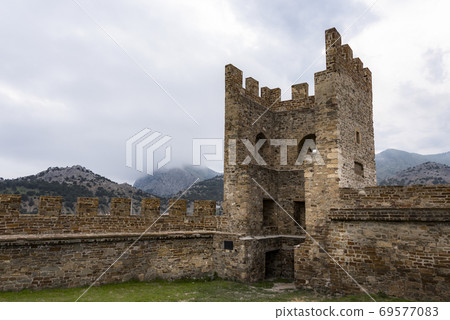 Tower and wall of Genoese fortress of 14th century in the Sudak bay on the Peninsula of Crimea on the background of mountains and cloudy sky 69577083