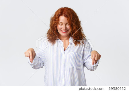 Beautiful middle-aged redhead woman in shirt looking and pointing fingers down with happy, cute smile. Concept of promo banners and advertisement, people reactions, white background 69580632