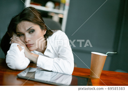 Tired or bored business woman relax at workplace. Young Caucasian woman rest at office table with coffee paper cup and digital tablet on it. Crisis concept. Tinted image 69585383