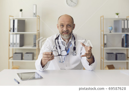 Senior experienced doctor speaks into camera with patient using gadget sitting in clinic office. Doctor online. 69587628