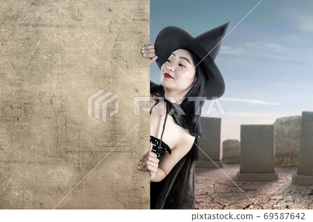 Asian witch woman with a hat standing with an empty board with a graveyard background Asian witch woman with a hat standing with an empty board with a graveyard background 69587642