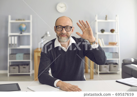 Waving old businessman smiling looking at the camera video call web camera gadget sitting in the apartment. Waving old businessman smiling looking at the camera video call web camera gadget sitting in the apartment. 69587659