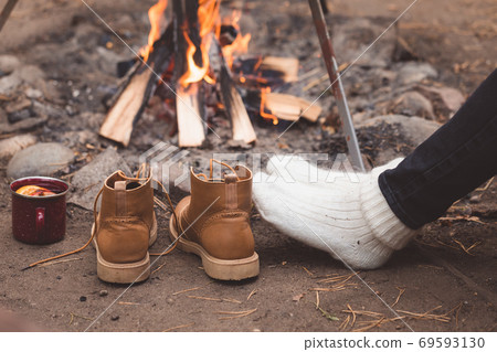 Spicy hot mulled wine in a mug, boots, legs with the knitted socks over the camp fire background Spicy hot mulled wine in a mug, boots, legs with the knitted socks over the camp fire background 69593130