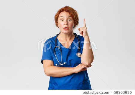 Overwhelmed redhead female doctor, middle-aged nurse in scrubs have suggestion, saying idea or plan, raising index finger eureka gesture with worried expression, standing grey background 69593930