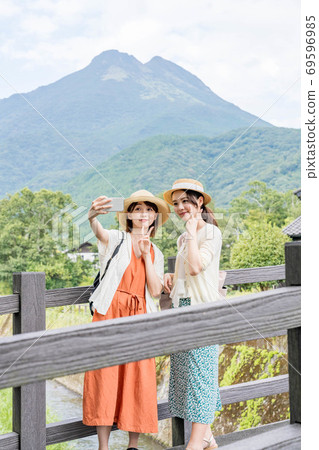 一名年輕女子和一位朋友在女孩出遊期間在由布山的背景下用智能手機射擊[9月] 69596985