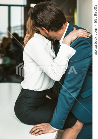 An office worker sits on a desk while a colleague kisses her neck. Passionate affair in the office workplace concept. High quality photo An office worker sits on a desk while a colleague kisses her neck. Passionate affair in the office workplace concept. High quality photo 69598005