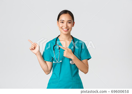 Covid-19, healthcare workers, pandemic concept. Smiling pretty asian doctor, nurse in scrubs and stethoscope, pointing fingers upper left corner, showing online clinic banner, white background Covid-19, healthcare workers, pandemic concept. Smiling pretty asian doctor, nurse in scrubs and stethoscope, pointing fingers upper left corner, showing online clinic banner, white background 69600658