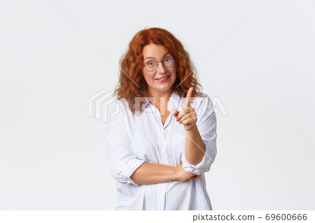 Portrait of middle-aged redhead woman in blouse and glasses explain something, pointing finger or shaking it in disapproval, teaching lesson, standing white background 69600666