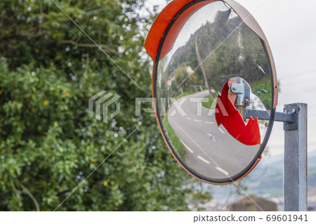Broken spherical road safety mirror with cracks and red plastic on a background of green foliage. Reflected asphalt road with white markings 69601941