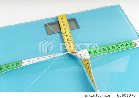Aquamarine floor scales wrapped in a multicolored centimeter on a white background. Tied in a knot. Tools for measuring weight and body parameters when dieting Aquamarine floor scales wrapped in a multicolored centimeter on a white background. Tied in a knot. Tools for measuring weight and body parameters when dieting 69601976