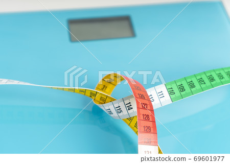 Aquamarine floor scales wrapped in a multicolored centimeter on a white background. Tied in a knot. Tools for controlling weight and body parameters when dieting Aquamarine floor scales wrapped in a multicolored centimeter on a white background. Tied in a knot. Tools for controlling weight and body parameters when dieting 69601977
