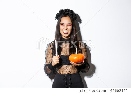 Image of asian evil witch in gothic lace dress and black wreath, laughing and grimacing, holding candle with pumpkin, celebrating halloween Image of asian evil witch in gothic lace dress and black wreath, laughing and grimacing, holding candle with pumpkin, celebrating halloween 69602121