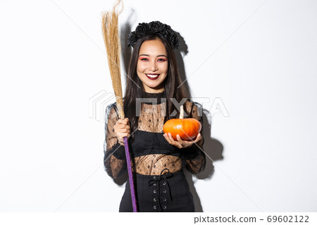 Image of beautiful asian woman dressed-up as a witch for halloween party, holding broom and pumpkin, standing over white background Image of beautiful asian woman dressed-up as a witch for halloween party, holding broom and pumpkin, standing over white background 69602122