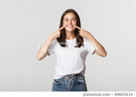Happy girl pointing fingers at mouth, showing white perfect teeth and beautiful smile Happy girl pointing fingers at mouth, showing white perfect teeth and beautiful smile 69603814