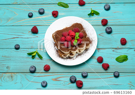 portion of Classic tiramisu dessert with raspberries and blueberries on blue wooden background 69613160