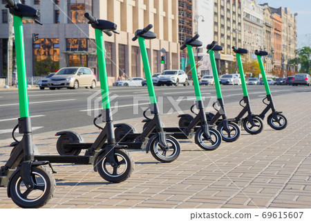 Eco transportation concept. Scooter rental in the city. Environment conservation. Eco friendly transportation. Vehicle rent service concept. Electric scooters in line on urban city background Eco transportation concept. Scooter rental in the city. Environment conservation. Eco friendly transportation. Vehicle rent service concept. Electric scooters in line on urban city background 69615607
