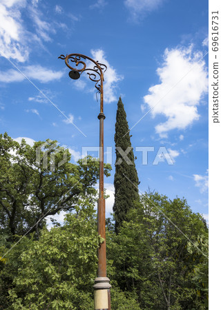 High rusty old pole with a lantern on the background of the cypress tree and cloudy sky High rusty old pole with a lantern on the background of the cypress tree and cloudy sky 69616731