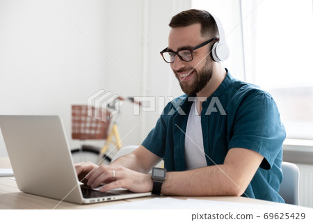 Smiling young man wearing headphones looking at laptop screen Smiling young man wearing headphones looking at laptop screen 69625429