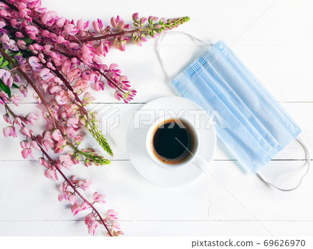 Cup of coffee, removed medical mask and pink flowers. Breakfast in the cafe. Modern reality and life concept. Blue surgical protective mask and white cup on white wooden background. Flat lay, top view Cup of coffee, removed medical mask and pink flowers. Breakfast in the cafe. Modern reality and life concept. Blue surgical protective mask and white cup on white wooden background. Flat lay, top view 69626970