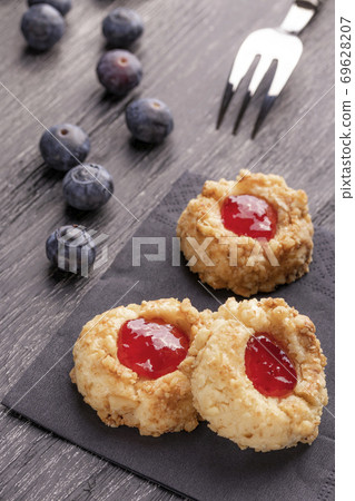 Cookies made from hazelnut shortcake with strawberry jam inside on a black napkin with blueberries and a patterned dessert fork on a dark background nearby. Cookies made from hazelnut shortcake with strawberry jam inside on a black napkin with blueberries and a patterned dessert fork on a dark background nearby. 69628207