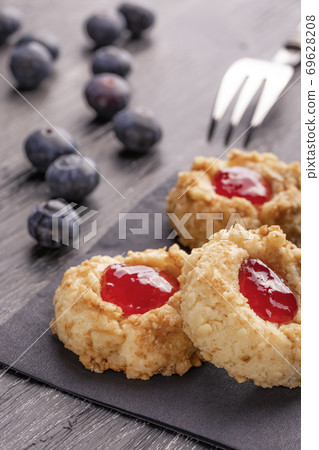 Cookies made from hazelnut shortcake with strawberry jam inside on a black napkin with blueberries and a patterned dessert fork on a dark background nearby. Cookies made from hazelnut shortcake with strawberry jam inside on a black napkin with blueberries and a patterned dessert fork on a dark background nearby. 69628208