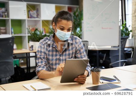 Back to office. Young woman, female office worker wearing protective face mask using digital tablet while sitting in the office Back to office. Young woman, female office worker wearing protective face mask using digital tablet while sitting in the office 69628791