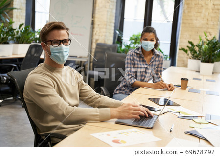 Working safely. Male and female office workers wearing medical protective masks sitting together at the table in the modern office and looking at camera 69628792