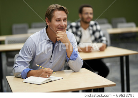Studying at work. Young happy man, male office worker sitting at desk in the office and smiling, listening to coach during corporate business training Studying at work. Young happy man, male office worker sitting at desk in the office and smiling, listening to coach during corporate business training 69628795