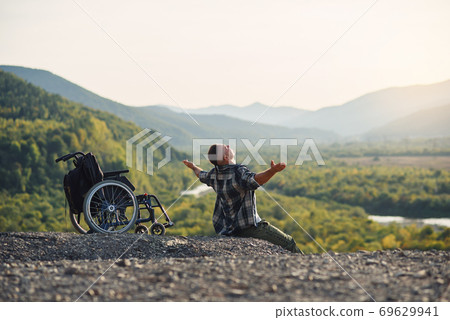 Young disabled man sitting on the mountain near wheelchair and raised hands up enjoying fresh air and nature beauty. Young disabled man sitting on the mountain near wheelchair and raised hands up enjoying fresh air and nature beauty. 69629941