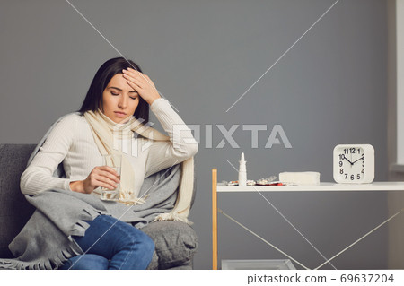 A sick girl with a fever looks at a thermometer while sitting on a sofa in the room. 69637204