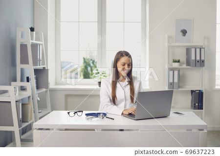 Smiling woman doctor sitting in medical clinic office with laptop and communicating online with somebody Smiling woman doctor sitting in medical clinic office with laptop and communicating online with somebody 69637223