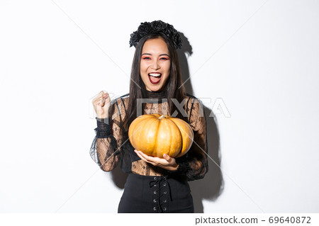 Image of excited beautiful asian woman celebrating halloween, wearing witch outfit and holding pumpkin, yelling with rejoice 69640872