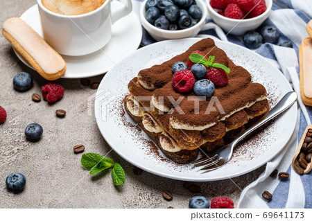 portion of Classic tiramisu dessert with raspberries, bluebedrries and cup of coffee on grey concrete background 69641173