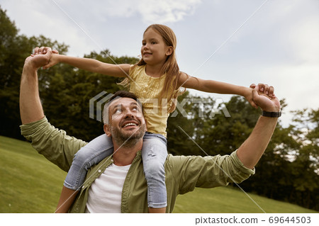Little excited girl playing with father in park, sitting on his shoulders and smiling, they are spending great time together and having fun Little excited girl playing with father in park, sitting on his shoulders and smiling, they are spending great time together and having fun 69644503