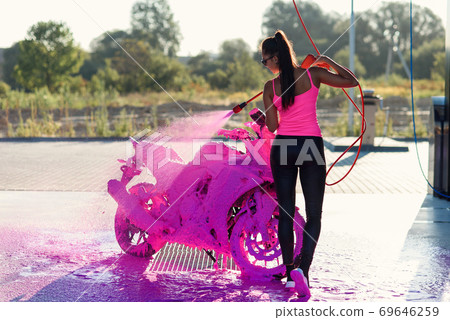 Beautiful stunning girl washes a motorcycle in self service carwash with high pressure foam jet. Beautiful stunning girl washes a motorcycle in self service carwash with high pressure foam jet. 69646259