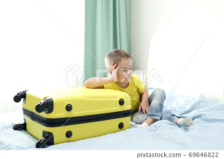blond boy is waiting for the holidays, sitting next to a yellow suitcase. A cute child with a suitcase is waiting for a taxi to go to the airport blond boy is waiting for the holidays, sitting next to a yellow suitcase. A cute child with a suitcase is waiting for a taxi to go to the airport 69646822
